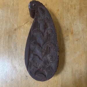 May include: A dark brown, teardrop-shaped wooden carving with a handle. The carving features a floral design with several rounded shapes and a crescent at the bottom. The wood has visible grain and a distressed appearance.