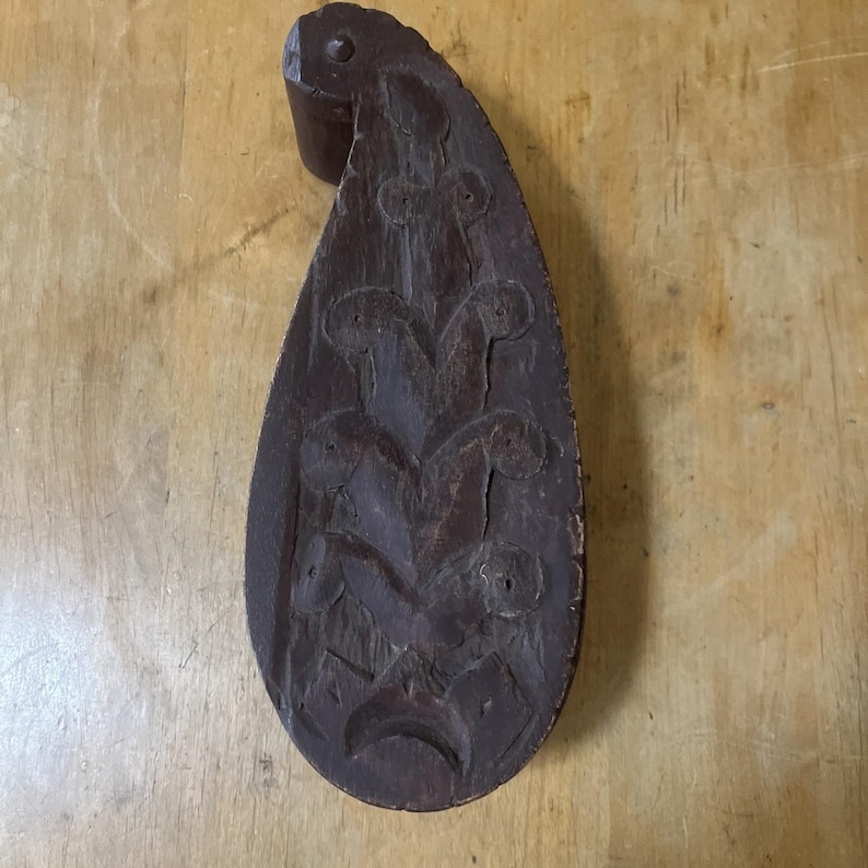 May include: A dark brown, teardrop-shaped wooden carving with a handle. The carving features a floral design with several rounded shapes and a crescent at the bottom. The wood has visible grain and a distressed appearance.
