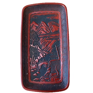 May include: A rectangular red lacquer tray with a black and red carved design of a mountain landscape. The tray has a decorative border with a repeating geometric pattern.