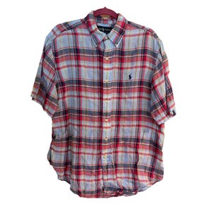 May include: Short-sleeved button-down shirt in a plaid pattern of red, blue, and yellow. The shirt has a button-down collar and a small embroidered logo on the chest. The shirt is hanging on a pink hanger.