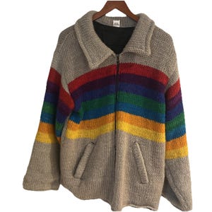 May include: A chunky knit zip-up sweater in a neutral beige color, featuring a vibrant rainbow design across the chest and sleeves. The sweater has a collared neckline, two front pockets, and is displayed on a wooden hanger.