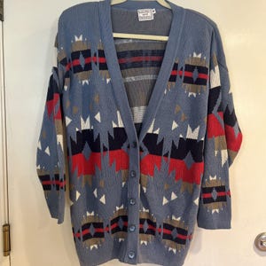 Könnte beinhalten: Blauer Cardigan-Pullover mit geometrischem Muster in Rot, Weiß, Marineblau und Braun. Der Pullover hat einen V-Ausschnitt, lange Ärmel und Knopfverschlüsse. Das Etikett lautet "Babette and Partners".