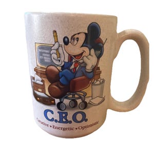 May include: A beige ceramic mug featuring a graphic of Mickey Mouse sitting in an office chair at a desk, holding a pencil and phone. The text reads C.E.O. Creative, Energetic, Optimistic. The mug has a textured surface and a single side handle.