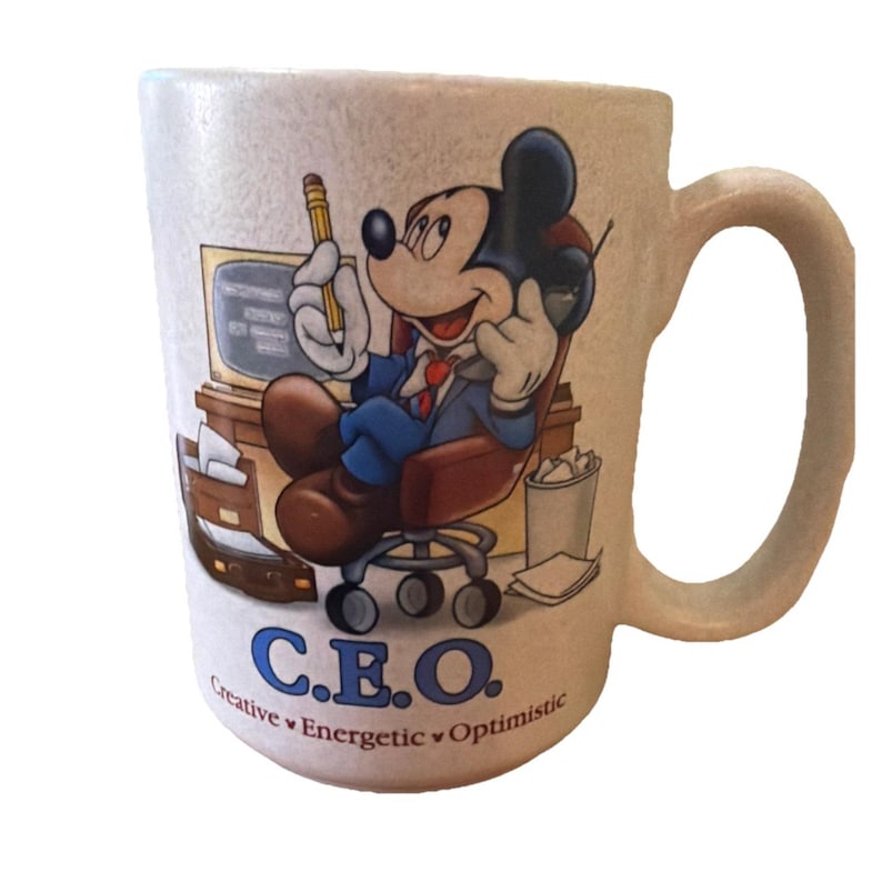 May include: A beige ceramic mug featuring a graphic of Mickey Mouse sitting in an office chair at a desk, holding a pencil and phone. The text reads C.E.O. Creative, Energetic, Optimistic. The mug has a textured surface and a single side handle.
