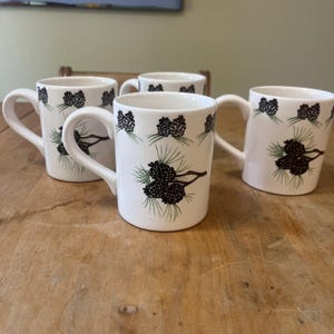May include: Four white ceramic mugs with a pine cone design. Each mug has a white handle and a repeating pattern of black pine cones and green pine needles. The mugs are arranged on a wooden table.