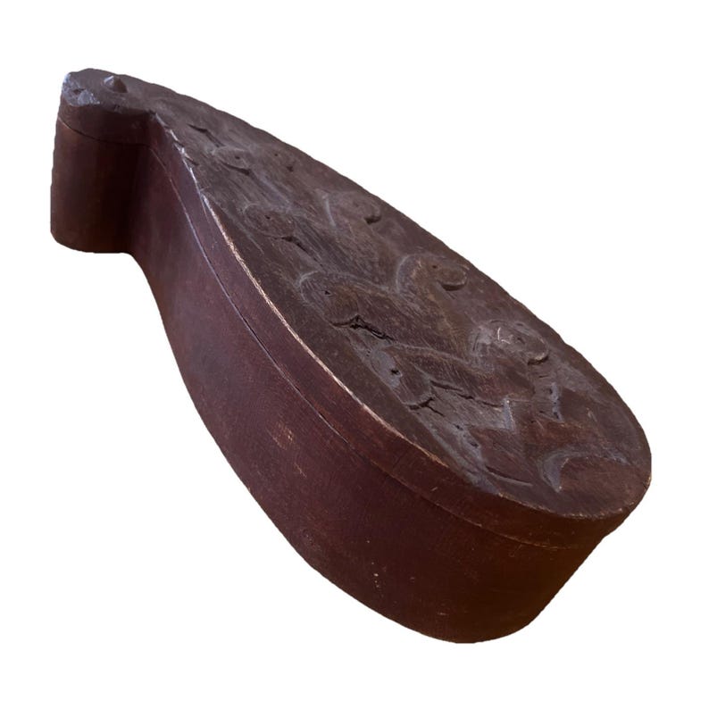 May include: A dark brown, wooden box with a curved, elongated shape. The lid features carved, abstract designs. The box has a rectangular base and a rounded end. The wood has a distressed appearance.