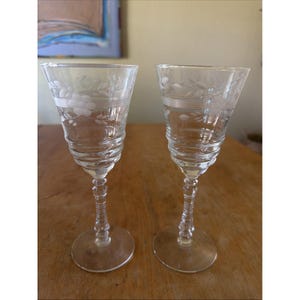 Op de afbeelding: Twee heldere glazen kelken met een delicaat geëtst bloemen- en bladpatroon rond de bovenrand. Elk glas heeft een geribbelde kom en een decoratieve, veelzijdige steel die uitloopt in een ronde voet, rechtopstaand geplaatst.