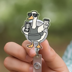 May include: A clear plastic badge reel with a cartoon goose wearing sunglasses, a grey apron, and holding an X-ray and a coffee cup. The reel has a silver retractable cord and a rectangular clip.