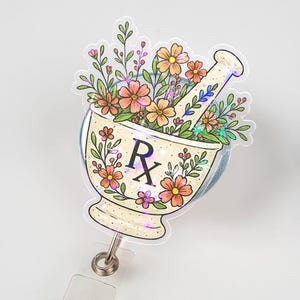 May include: A badge reel featuring a mortar and pestle design filled with flowers in shades of orange, pink, and green. The mortar has the letters "Rx" on it. The reel has a clear plastic strap and a metal clip.