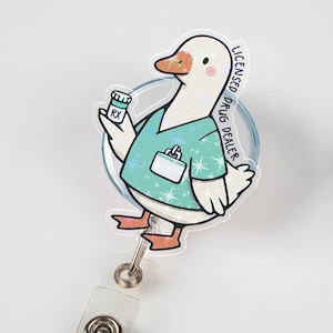 May include: A badge reel featuring a cartoon goose in teal scrubs, holding a prescription bottle. The text "LICENSED DRUG DEALER" is visible. The goose has an orange beak and feet, and the reel has a clear plastic clip.