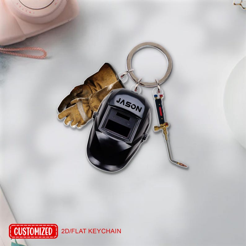 May include: A keychain featuring a black welding helmet with the name "JASON", brown work gloves, and a miniature welding torch. The keychain is attached to a silver ring. The text "CUSTOMIZED" and "2D/FLAT KEYCHAIN" is visible.
