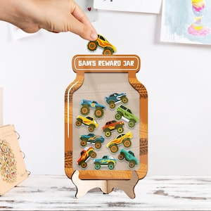 Peut inclure: Un pot de récompenses en bois avec le texte "SAM'S REWARD JAR". Le pot est rempli de camions monstres jouets colorés. Une main place un camion monstre jaune et orange au-dessus.