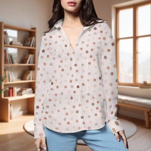 May include: A button-down shirt with a white background and a pattern of various-sized brown, tan, and gray polka dots. The long-sleeved shirt has a collared neckline and two small pockets. The model is wearing blue jeans.
