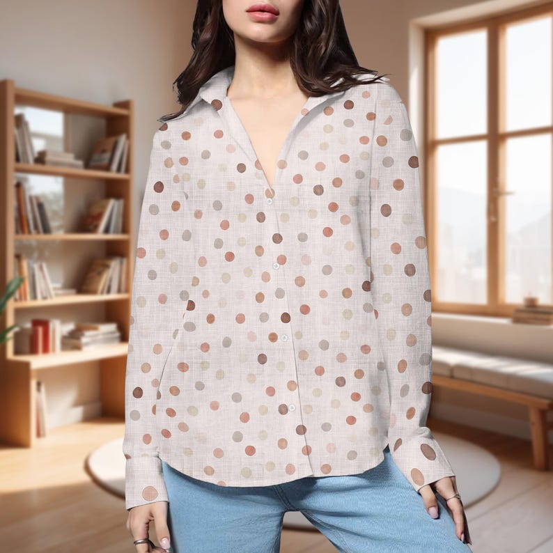 May include: A button-down shirt with a white background and a pattern of various-sized brown, tan, and gray polka dots. The long-sleeved shirt has a collared neckline and two small pockets. The model is wearing blue jeans.