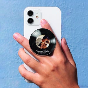 Op de afbeelding: Een zwarte telefoonhouder in de vorm van een vinylplaat, met een foto van een stel. De houder is bevestigd aan een witte smartphone. De tekst "Sophia and Brandon" en de datum "12.20.2020" staan op de houder.