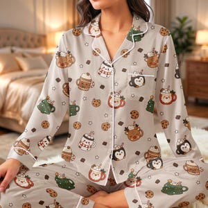 Op de afbeelding: Een lichtbeige pyjamaset met lange mouwen, knoopsluiting en bijpassende broek, voorzien van een herhalend patroon van getekende rendieren, pinguïns, kerstbomen en mokken warme chocolademelk met slagroom en kleine bruine chocoladestukjes.