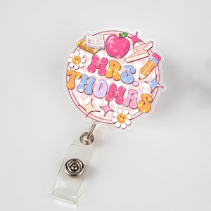 May include: A vibrant badge reel featuring the name "MRS. THOMAS" in a retro font. The design incorporates an apple, pencil, book, and daisies. The reel has a clear plastic strap and a metal clip.