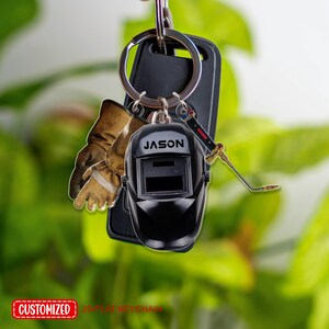 May include: A black welding helmet keychain with the name "JASON" printed on it. The keychain also includes a miniature welding torch and a pair of brown work gloves. The keychain is attached to a silver keyring.