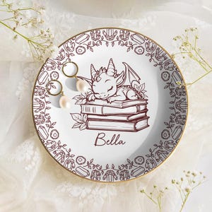 May include: White ceramic trinket dish with a gold rim, featuring a brown illustration of a sleeping dragon on books. The dish is decorated with a border of crystals and other shapes. A pair of gold earrings with pearls rests on the dish.