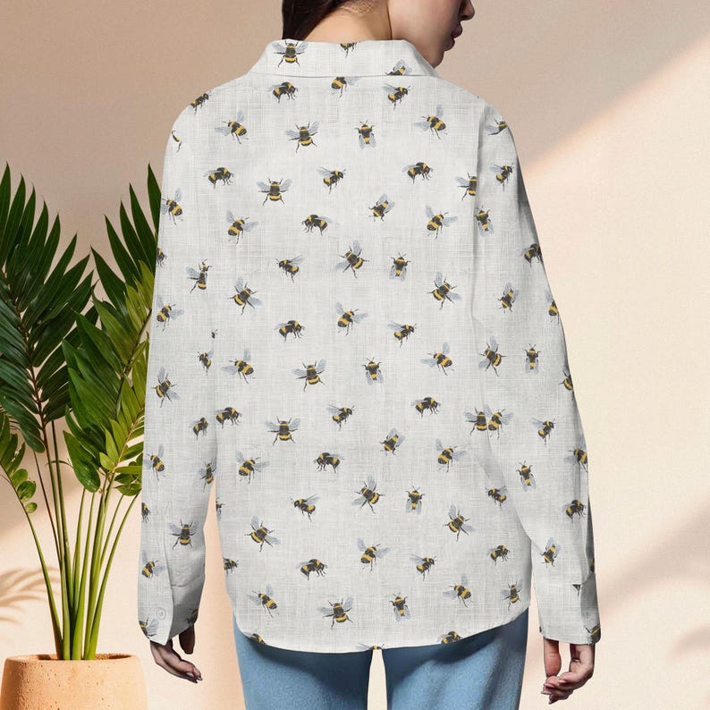 May include: A white button-down shirt with a repeating pattern of yellow and black bees. The long-sleeved shirt is made of a light-colored fabric and has a classic collar and buttoned cuffs. The bees are scattered across the shirt.