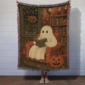 Puede incluir: Una manta tejida que presenta un fantasma de dibujos animados leyendo un libro en un ambiente otoñal y acogedor. El diseño incluye una calabaza, una ventana con una linterna de calabaza y una estantería. La paleta de colores es cálida, con tonos marrones, naranjas y verdes.