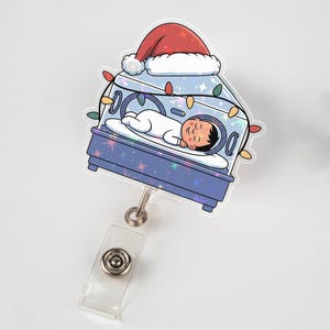 May include: A badge reel with a festive design. It shows a baby in an incubator wearing a Santa hat, with colorful lights. The baby is dressed in white. The reel has a clear plastic clip and a silver metal retractable cord.