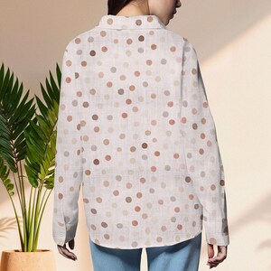 May include: A white button-down shirt with a pattern of various-sized polka dots in shades of brown, tan, and gray. The long-sleeved shirt is made of a lightweight fabric and has a classic collar and buttoned cuffs. The shirt is worn with blue jeans.