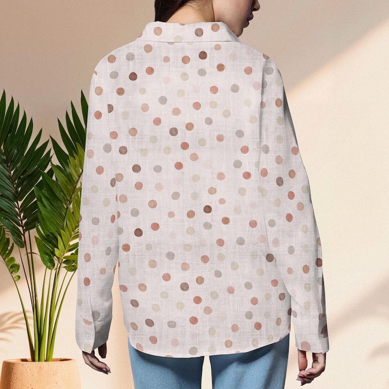 May include: A white button-down shirt with a pattern of various-sized polka dots in shades of brown, tan, and gray. The long-sleeved shirt is made of a lightweight fabric and has a classic collar and buttoned cuffs. The shirt is worn with blue jeans.