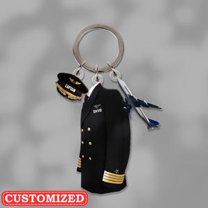 Könnte beinhalten: Ein Schlüsselanhänger mit einer Pilotenuniform, einer Mütze und einem Flugzeuganhänger. Die schwarze Uniform hat goldene Knöpfe und Streifen. Auf der Mütze steht "CAPTAIN" und der Name "DAVID" steht auf der Uniform. Das Wort "CUSTOMIZED" steht auf einem roten Banner.