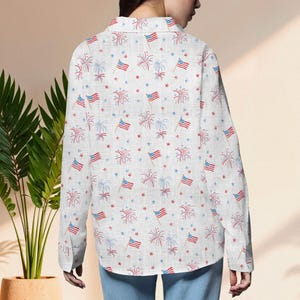 K&ouml;nnte beinhalten: Wei&szlig;es Button-Down-Hemd mit einem sich wiederholenden Muster aus amerikanischen Flaggen, roten und blauen Feuerwerken sowie roten und blauen Sternen. Das Hemd hat lange &Auml;rmel und einen Kragen. Der Stoff scheint eine leichte Baumwolle oder Leinen zu sein.