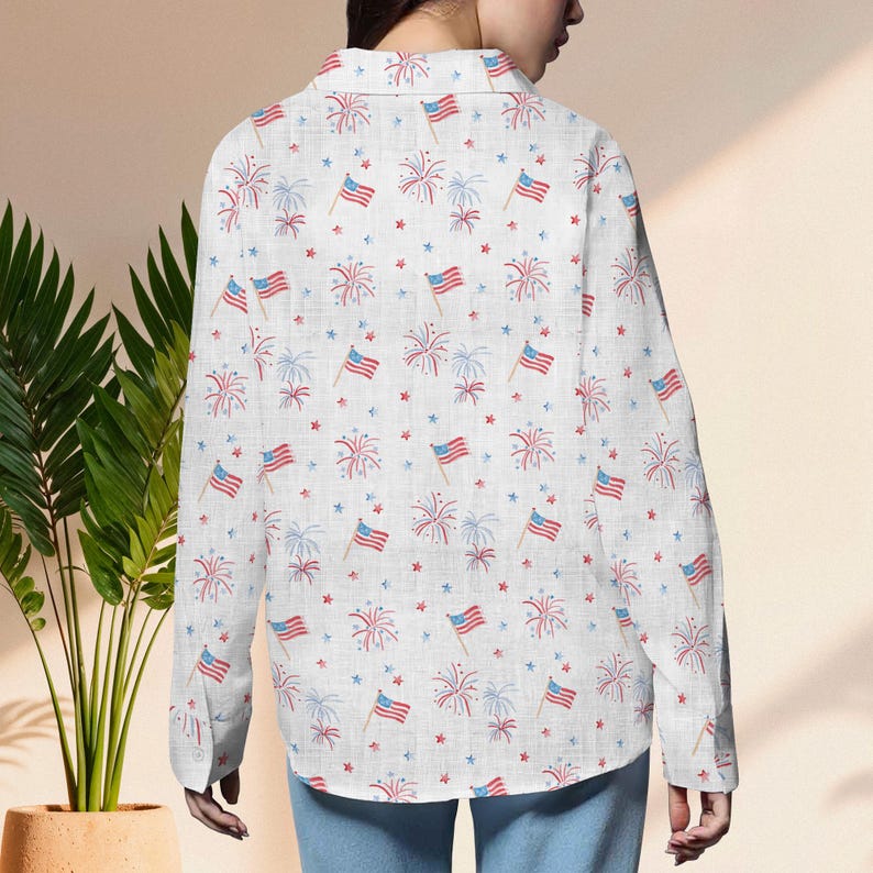 K&ouml;nnte beinhalten: Wei&szlig;es Button-Down-Hemd mit einem sich wiederholenden Muster aus amerikanischen Flaggen, roten und blauen Feuerwerken sowie roten und blauen Sternen. Das Hemd hat lange &Auml;rmel und einen Kragen. Der Stoff scheint eine leichte Baumwolle oder Leinen zu sein.