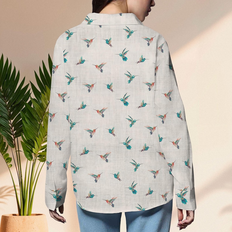 May include: A light-colored button-down shirt featuring a repeating pattern of hummingbirds in flight. The birds have teal, orange, and green accents. The shirt has long sleeves and a collared neckline. The shirt is made of a lightweight fabric.