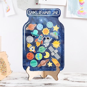 Peut inclure: Un pot à récompenses en bois sur le thème de l'espace, avec des planètes, des fusées et un astronaute sur fond étoilé. Le pot est étiqueté "SAM'S REWARD JAR" et est soutenu par un support en bois.