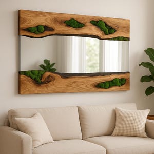 Puede incluir: Un espejo rectangular con marco de madera natural. El marco tiene formas orgánicas con inserciones de musgo verde. El espejo refleja una ventana y una planta. El espejo está colgado sobre un sofá beige.