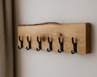 Puede incluir: Un perchero de madera con siete ganchos de metal negro. El perchero está hecho de una pieza rectangular de madera de color claro con un borde natural en la parte superior. Los ganchos están espaciados uniformemente a lo largo del borde inferior.