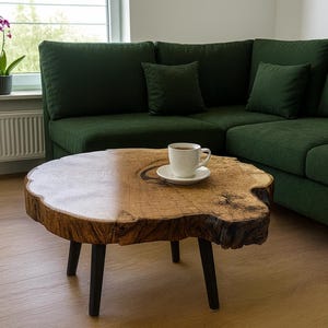 Peut inclure: Une table basse ronde en bois avec un bord naturel et trois pieds noirs. Une tasse à café blanche et une soucoupe sont posées sur la table. Un canapé vert foncé est en arrière-plan.