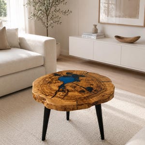 Op de afbeelding: Ronde houten salontafel met een natuurlijke houtnerf afwerking en een blauwe harsinleg. De tafel heeft drie zwarte poten en is ongeveer 61 cm in diameter.