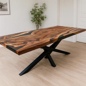 Peut inclure: Une table de salle à manger rectangulaire en bois avec une incrustation de résine époxy foncée. La table a un fini grain de bois naturel et repose sur un piètement en métal noir en forme de X. La table mesure environ 183 cm de long.