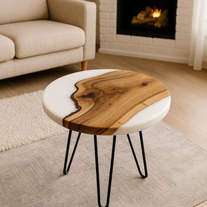 Puede incluir: Mesa auxiliar redonda con tapa de resina blanca e incrustación de madera natural. La mesa tiene patas de horquilla negras. La veta de la madera es visible en el centro de la mesa. La mesa tiene unos 51 cm de diámetro.