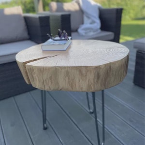 May include: A round, light-coloured wooden side table with a natural wood grain finish. The table has three metal hairpin legs. A book and glasses rest on the table's surface. The table is approximately 61 cm in diameter.