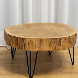 Puede incluir: Mesa de centro redonda de madera natural clara. Tiene tres patas de horquilla de metal negro. La veta de la madera es visible en la tapa y los lados. El diámetro es de aproximadamente 76 cm.