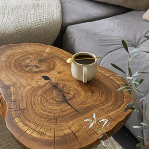 Puede incluir: Una mesa de centro de madera maciza con bordes naturales, una taza de café y una cucharita. La mesa tiene un patrón de vetas de madera natural y está rodeada por un sofá gris y plantas decorativas. La mesa tiene unos 76 cm de diámetro.
