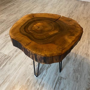 Puede incluir: Una mesa auxiliar redonda de madera con borde natural y acabado marrón oscuro. La mesa está sostenida por tres patas de horquilla negras. La veta de la madera es visible y la mesa mide aproximadamente 50 cm de diámetro.