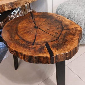 Puede incluir: Mesa de centro redonda de madera con borde natural y acabado marrón oscuro. Tiene tres patas de metal negro. La superficie presenta un patrón de vetas de madera único y detalles oscuros. Diámetro aproximado de 60 cm.