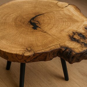 May include: A round wooden coffee table with a natural, unfinished wood top. The table has a dark, black resin-filled crack and is supported by three black metal legs. The wood grain is visible, and the table is approximately 30 inches in diameter.