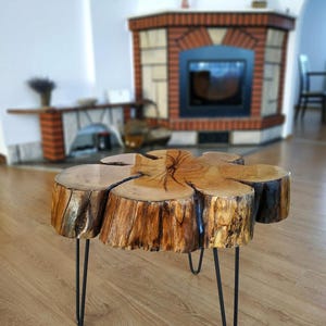 Op de afbeelding: Een rustieke salontafel gemaakt van een dwarsdoorsnede van een boomstam. Het lichtbruine hout heeft zichtbare nerven en donkerbruine randen. Hij staat op drie zwarte haarspeldpoten. Diameter: ongeveer 75 cm.