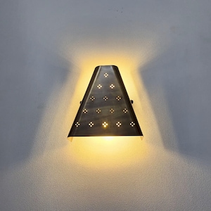 “Vintage brass wall sconce turned on, trapezoid shade with floral perforated cutouts, warm yellow glow on grey wall, handmade modern wall light.”