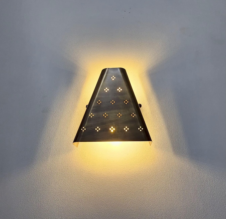 “Vintage brass wall sconce turned on, trapezoid shade with floral perforated cutouts, warm yellow glow on grey wall, handmade modern wall light.”