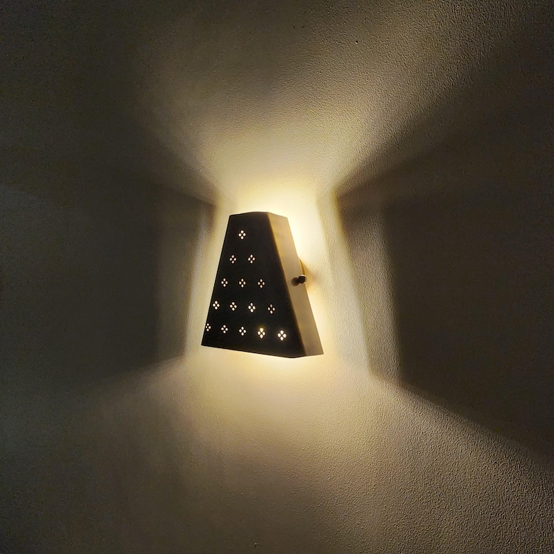 “Handmade vintage brass wall sconce lit up on wall, trapezoid shade with perforated cutout pattern, warm ambient up and down light effect.”