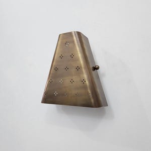 May include: A brass-colored wall sconce with a trapezoidal shape. The sconce has a series of small, evenly spaced holes and a small, round knob on the side. It is mounted on a white wall.